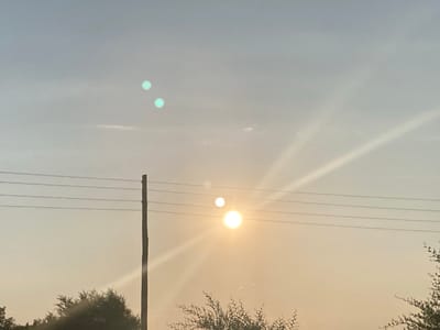 A power pole stands to the left of the frame, with four parallel wires stretching across it. The sun is low in the sky, casting a warm glow and creating lens flares. Two of these flares appear as bright, circular orbs, with one significantly larger and more intense than the other. There are also two smaller, teal-colored orbs higher in the sky, likely due to lens artifacting. The background consists of a hazy sky with faint clouds. The bottom of the frame shows the dark silhouettes of trees and foliage. The scene is set during the day, under an overcast or hazy sky, with the sun being a prominent element. The location is specified as the city of Rakity, Russia.