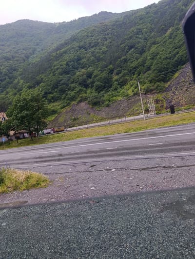 This image depicts a roadside scene with a lush, green mountainside in the background. The mountain is densely covered in trees, with a rocky, barren slope visible on the right side. A paved road with white lane markings runs across the foreground, appearing wet, suggesting recent rain. The shoulder of the road is a mix of gravel and sparse vegetation.

Behind a metal fence bordering the road, there are several buildings with visible windows, some appearing to be of a residential or industrial nature. Colorful trucks are parked near these buildings, and a blue road sign is also present. A power line pole with visible wires stretches across the scene, and further up the mountain, there's a white metal structure, possibly related to infrastructure. The sky is overcast and a light gray, indicating a cloudy or possibly foggy day. The overall mood is serene and natural, with a touch of human activity along the roadside. The location is identified as Nizhnii Lars, Russia.