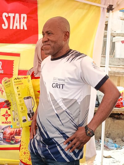 This media file captures a portrait of a man standing outdoors, possibly at a market or event in Tarauni, Nigeria. He is casually dressed in a white t-shirt with a distinctive blue and grey abstract design and black trim. On his left wrist, he wears a black wristwatch.

The man is positioned in front of a stall or tent with a bright yellow backdrop, and a red banner with "STAR" printed in large white letters is visible to his left. To his right, a white tent pole and some construction materials are present, suggesting an outdoor setting.

In the foreground, several products are displayed, including a yellow box for a "Mewe Blender & Grinder" and a red package featuring "MAGGI" and an image of a roasted chicken. These items suggest the man might be involved in a promotional event or is a vendor.

The man's facial expression is a slight smile and he is looking off to his right, giving the impression he is engaged in a conversation or observing something. The lighting suggests it is daytime. The overall scene conveys a lively atmosphere, typical of a market or community gathering.