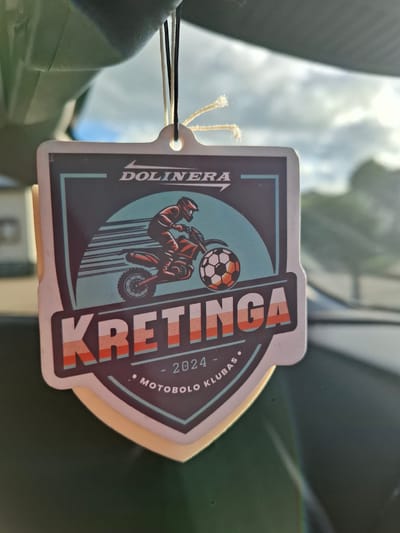 A close-up, slightly angled shot shows a car air freshener hanging from a car's interior. The air freshener is a shield-shaped cardboard tag with a graphic design. The design features a dirt bike rider in action with the word "DOLINERA" above and "KRETINGA" below in large, stylized letters. A soccer ball is depicted near the dirt bike. The bottom of the tag has "2024 - MOTOBOLO KLUBAS" written in a circular pattern. The air freshener is suspended by two strings, one black and one off-white, which are tied together at the top. The background is blurred, showing the sky with clouds and hints of trees and buildings, suggesting an outdoor environment viewed through the car's windshield. The interior of the car is visible in the foreground, with a dark green dashboard and a reflection of light on the windshield. The image is well-lit, with the sun casting a warm glow. The location is stated to be in Huntingdonshire, United Kingdom, although no specific visual cues in the image confirm this.