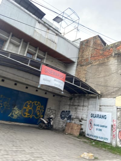The image shows a narrow alleyway in Margahayu, Indonesia (latitude: -6.965, longitude: 107.577). The alley is lined with brick walls and buildings on both sides, with a metal awning overhead. A blue metal gate with graffiti painted on it can be seen in the background. There is a motorbike parked in the alley, facing away from the camera. On the right wall, there is a sign that reads "DILARANG BUANG SAMPAH DI SINI" in white text on a blue background. The sign is attached to a brick wall, and the wall is covered in a layer of dust and dirt. There are several power lines running across the alleyway.