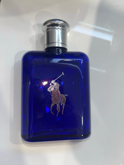 The image displays a deep blue, rectangular glass bottle, likely for cologne or perfume, positioned horizontally on a clean, white, and mildly reflective surface. The bottle features prominent rounded edges and a polished silver cap on its left side. A detailed silver emblem of a polo player on horseback, in mid-swing with a mallet, is affixed to the front of the blue bottle. The emblem shows a textured finish, contrasting with the smooth glass.
Partial text, "RALPH L", is visible on the silver band connecting the cap to the bottle, suggesting the brand name Ralph Lauren. The translucent blue glass exhibits various internal light reflections and refractions, as well as some minor surface dust and streaks. The white background surface also shows subtle reflections of light sources and a few faint marks. The scene is a still life photograph of the product, captured indoors under what appears to be artificial lighting. No specific activity or event is taking place.