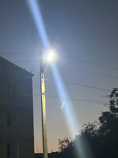 A street lamp with a bright, horizontal light fixture illuminates the sky with a strong beam, casting a long, blueish-white glare downwards. The lamp is mounted on a tall, light-colored utility pole, which is positioned to the right of a dark, multi-story building. The building has at least two visible windows on its facade. Power lines run horizontally across the scene, strung between poles and the building. To the right of the utility pole and extending into the lower right quadrant of the image is a cluster of dark tree branches and foliage. The sky is a dusky gradient, transitioning from a lighter tone near the horizon to a deeper blue above, suggesting it is either early morning or late evening. The setting appears to be an urban or suburban environment in Şamaldı-Say, Kyrgyzstan, characterized by the presence of buildings, utility poles, and street lighting. No people are visible in the frame.