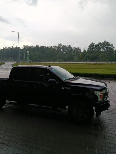 The image depicts a gas station forecourt and an adjacent highway under an overcast sky in Yosypivka, Ukraine. A dark-colored Ford F-150 XLT pickup truck is parked on the wet, dark grey paver-covered surface, facing toward the right foreground. Its surfaces appear wet, consistent with recent or ongoing precipitation. In the midground, a tall green sign displays a "УОГ" (UOG) logo at the top and electronic price panels for fuel types including A-95, ДП (Diesel), A-92, and ГАЗ (LPG). Behind the gas station area, a multi-lane highway is visible, separated by a grassy embankment and a metal guardrail. A dense treeline composed of green deciduous trees forms the background. The sky is uniformly light grey and overcast. Multiple tall streetlights are present within the scene. No individuals are visible, and no specific actions are depicted beyond the parked vehicle. The entire outdoor environment appears wet.