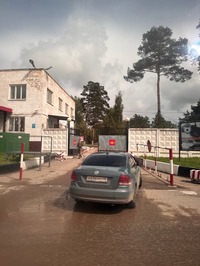 The image captures an outdoor scene at what appears to be a security checkpoint or entrance to a military facility, under an overcast sky during daytime. The ground is wet, suggesting recent rain.

In the foreground, a grey Volkswagen sedan, identifiable by its logo and taillights, is positioned on a wet asphalt surface. Its Russian license plate, "A636PC 18", is clearly visible. Red and white striped barriers, possibly boom gates or traffic markers, are prominent near the vehicle and across the entry point.

To the left of the scene stands a two-story building constructed from white brick, featuring multiple windows and two street lights mounted on its side. A green metal structure is located in front of the building.

Central to the image is a dark metal gate, prominently displaying a red shield with a yellow double-headed eagle holding a sword and scepter—the emblem of the Russian Ministry of Defense. Behind this gate, a textured white concrete block fence forms a perimeter. A person in a military uniform, likely a guard, is visible standing near the gate on the far side. A smaller sign with the same Ministry of Defense emblem is also present further inside the gate.

Above the fence, a black and white sign features a military insignia, including a star and what appears to be a hammer and sickle emblem, alongside partially visible Russian text that reads "39 гвардейская..." (39th Guards...). Tall pine trees are scattered throughout the background, contributing to the setting. A bright glare, likely the sun, is visible through the clouds and trees in the upper left. The overall scene suggests a functional and secure entry point.
