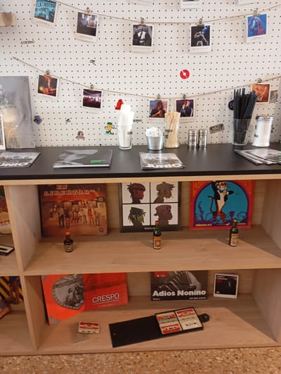 The image depicts a close-up, eye-level view of a shelf display, likely in a shop or private collection. The background is a white pegboard with black dots, strung with a thin twine from which various small, clipped photographs are suspended. These photographs appear to be of musicians or bands performing, some in black and white and others in color, clipped by small metal clips.
On the main shelf, there are several items. To the left, partially visible, is a stack of items including what looks like album covers or magazines. To the right of these, there is a black square object, possibly a book or record, with the word "JUNIO" visible. Further to the right is a stack of what appears to be records or CDs. In the center of the shelf is a black flat surface holding two more clipped items, one a book with "FEDERICO MOURA" printed on it.
Below this main shelf, a second shelf displays a variety of items. On the left is a large square item with the text "EN LIBERTAD" prominently displayed and a group of people pictured. To its right is a framed item featuring cartoon characters. Further right is a record with a tiger graphic and the text "volumen nº 5". On the far right, another item is visible.
On the second shelf, in front of the displayed items, are two small dark bottles, appearing to be miniature liquor bottles.
The bottom shelf, visible at the very bottom of the image, shows more items, including what appears to be an orange square item with text, and further to the right, a black open case containing four cassette tapes.
The overall impression is one of a curated display of music memorabilia. The mention of "La Plata, Argentina" suggests this collection might be related to the local music scene of that city. The specific items like "EN LIBERTAD" and "FEDERICO MOURA" hint at potential connections to Argentine artists and music history.