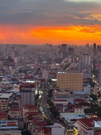 This aerial view captures a dense urban landscape of Khan Mean Chey, Cambodia, under a vibrant, cloud-filled sunset. The sky is dominated by a gradient of fiery orange and yellow, transitioning into dusky gray clouds. Below the dramatic sky, a sprawling cityscape unfolds, characterized by a multitude of buildings of varying heights and architectural styles. Many structures feature red-tiled roofs, contrasting with the concrete and glass of taller modern buildings. The streets are visible, illuminated by streetlights and the glow of vehicle headlights, indicating evening or early night. Traffic appears moderate. Trees are interspersed throughout the city, especially along the roadways. A prominent, light-yellow, multi-story building with a grid-like facade stands out in the mid-ground. Another tall, slender building with a patterned grey facade is also visible. The overall impression is one of a bustling urban center at the close of day, with the natural spectacle of the sunset providing a dramatic backdrop. No individuals are clearly discernible at this distance and resolution.