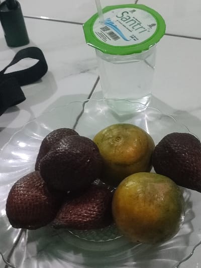 A clear glass bowl sits on a white tiled surface, containing 5 fruits. The bowl is filled with 4 brown salak fruits, with rough, textured skin and 1 yellow citrus fruit. There are two similar yellow citrus fruits, one partially obscured by the bowl's rim. A clear glass with a green lid featuring the word "SANTri" in white lettering sits beside the bowl. The glass is filled with clear liquid, likely water. The scene appears to be inside a house or a room. The location is Kecamatan Pandaan, Indonesia, based on coordinates (latitude: -7.668, longitude: 112.724).