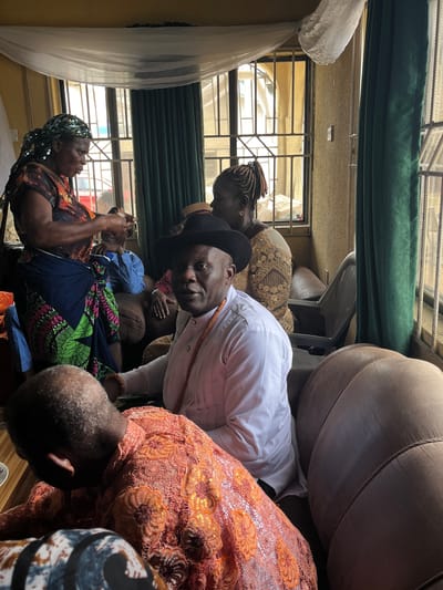 A low-angle medium shot shows a group of people in Warri, Nigeria. A man in a white shirt and black hat faces the camera with a slightly tilted head. A woman in a patterned dress and headscarf lies in a seated position. The rest of the people are obscured by the foreground. A window with bars is visible in the background. There is a dark green valance at the top of the window. A sofa is visible.