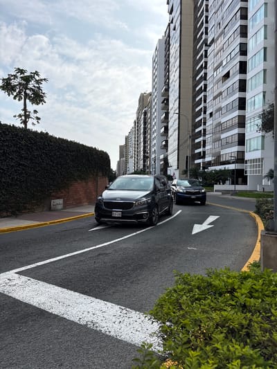 The image captures a street scene in San Isidro, Peru, characterized by modern high-rise buildings and a tree-lined road. A dark gray Kia Carnival minivan is driving on the road, with its license plate reading "BCH-690". A blue SUV is visible further down the road, also in motion. The road has white directional arrows and a yellow border. On the left side of the road, a dense green hedge covers a brick wall, with a slender tree rising above it. The sky is partly cloudy.