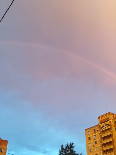The image captures a serene sky in Shchyolkovo, Russia, displaying a faint rainbow arching across the pale pink and blue clouds. The lower portion of the frame reveals the tops of two residential buildings. The building on the right is taller and appears to be made of a yellow-orange material, featuring multiple windows, balconies, and decorative architectural elements. The building on the left is partially visible and seems to be constructed with a similar material. A dark wire stretches across the sky, and a few trees are visible at the bottom left. The overall scene is a peaceful urban landscape under a colorful sky.