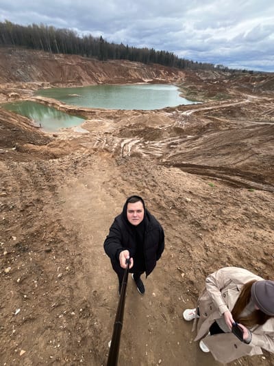 The image shows a man taking a selfie with a selfie stick. He is standing on a dirt cliff edge overlooking a quarry lake in Ivlevo, Russia. The quarry has steep, brown dirt walls and a lake at the bottom. Another person wearing a beige coat and a grey hat is standing nearby and using a cell phone. The sky is cloudy.