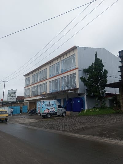 A three-story building with numerous windows faces the viewer on an overcast day. A delivery truck with child-themed graphics is parked in front, near two large blue water tanks. A motorcycle is parked nearby. To the left, a billboard advertises "Galaxy Resort." A yellow vehicle is partially visible on the left, and a person is seen near a blue fence. A tall, slender tree stands next to the building. The street in the foreground is wet.