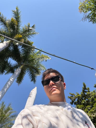 A low-angle selfie features a person wearing sunglasses and a white sweater. The backdrop is a clear blue sky dotted with palm trees and green foliage. A thin pole extends upwards across the sky. The person's face is partially obscured by the sunglasses and is angled downwards towards the camera. The bright lighting suggests a sunny day.