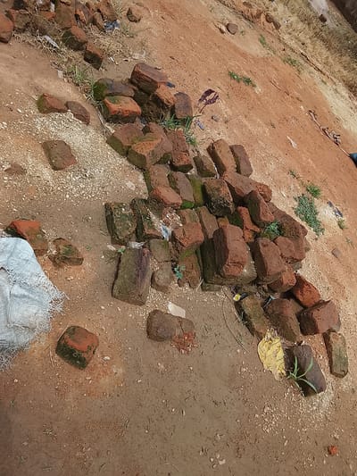 This media file depicts a pile of weathered, red bricks scattered on reddish-brown dirt and gravel. Some of the bricks show signs of moss or lichen growth, indicating they have been exposed to the elements. Small patches of green grass and dry weeds are interspersed among the bricks and on the surrounding ground. In the lower left corner, a portion of a torn, white woven bag is visible, suggesting a construction or storage area. The scene is outdoors, under daylight, with no specific indication of weather conditions beyond what can be inferred from the dry ground and grass. The overall impression is one of disarray, possibly a construction site, a demolition site, or simply discarded building materials. There is no discernible activity or event taking place, and no people or text are visible in the image. The location is identified as Sunkani, Nigeria, which is consistent with the earthy tones and vegetation.