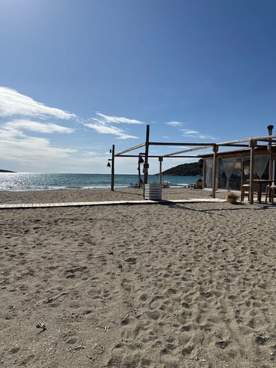 A bright, sunny beach scene in Anatoliki Attiki, Greece, is depicted under a clear blue sky. The foreground is dominated by a wide expanse of dark greyish-brown sand, textured with numerous footprints and scattered small green plants. A narrow, light-colored wooden boardwalk or pathway runs along the edge of the sand, separating it from the water.

In the mid-ground, a rustic wooden structure stands near the waterline. This appears to be a beach bar or shelter, featuring exposed timber beams, a simple roof, and transparent plastic curtains or windows. A white wooden pallet or crate is visible next to the structure.

Beyond the wooden building, the sparkling blue-green sea extends to the horizon. Sunlight glints off the water's surface, creating bright reflections. A distant dark landmass, likely a hill or mountain, lines the far shore. The sky above is a vibrant blue, adorned with a few elongated white clouds.

The image conveys a sense of peacefulness and quiet, with no people visible on the beach or in the water. The bright sunlight suggests it is daytime, either morning or early afternoon, with calm and clear weather conditions. No discernible text is present in the scene.