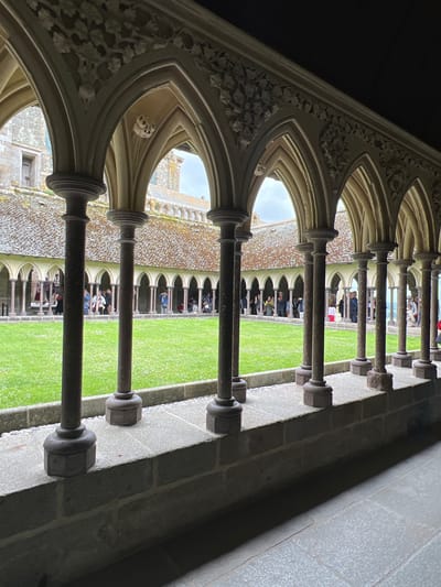The image depicts a cloister walkway with people observing a grassy courtyard. The walkway is lined with stone columns and arched openings, with intricate carvings visible on the interior arches. Sunlight filters through the arches, illuminating the green grass and casting shadows.

The subjects are groups of people, some standing and others seated, looking out into the courtyard. They appear to be tourists or visitors. The objects include the architectural elements of the cloister, such as the stone pillars, arches, and carved details, as well as the people themselves. The scene is an exterior cloister of a historical building, likely a monastery or abbey, given the architectural style.

The activity taking place is observation of the courtyard and the surrounding architecture. It appears to be a daytime scene, with bright lighting. The weather seems fair, as indicated by the clear sky visible through some of the arches. The location cues, such as the architectural style, strongly suggest Le Mont-Saint-Michel, France. The mood appears calm and contemplative, consistent with a visit to a historical and religious site. There is no visible text in the image.