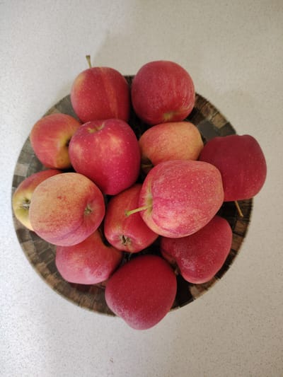 A top-down, medium shot shows a collection of approximately a dozen red and yellow apples nestled within a dark, woven bowl. The apples are the primary subjects, exhibiting varying degrees of red and yellow coloration and speckling. The bowl itself appears to be made of dark, textured material, possibly wood or a similar natural fiber, and sits on a speckled, light-colored surface. The background is a continuation of this light-colored surface, with some subtle shadows cast by the objects. There are no people or discernible actions taking place. The scene is static and focuses solely on the arrangement of the fruit in the bowl. The location is not specified within the image content.