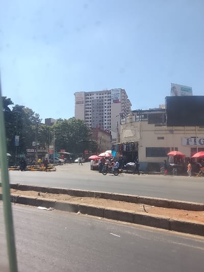 A bustling street scene under a clear sky features pedestrians and motorcycles near crosswalks and red parasols. A tall building displays "Park Inn" on its side, while a sign reading "FIG..." is visible on a lower structure. Street vendors are present, selling clothing. The foreground includes a curb separating the street from a dirt shoulder. The photo appears to be taken from a moving vehicle.