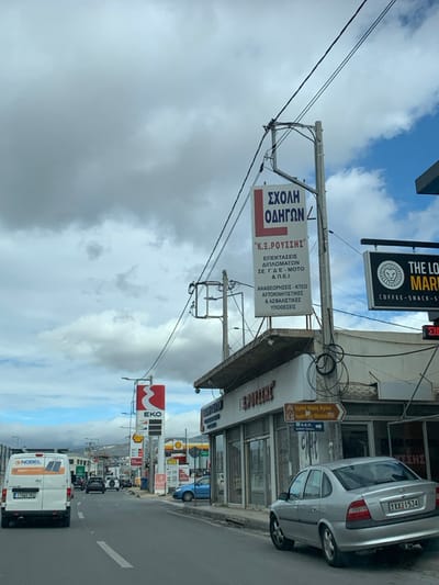 This image captures a street scene in Agia Paraskevi, Greece, under a partially cloudy sky during daylight hours. The setting is an urban street lined with commercial buildings.

Prominently featured is a large white sign for "ΣΧΟΛΗ ΟΔΗΓΩΝ Κ.Ξ. ΡΟΥΣΣΗΣ" (K.X. ROUSSIS Driving School), detailing services like "ΕΠΕΚΤΑΣΕΙΣ ΔΙΠΛΩΜΑΤΩΝ" (license extensions) for various vehicle categories (C-D-E, Moto, P.E.I.), "ΑΝΑΘΕΩΡΗΣΕΙΣ - ΚΤΕΟ" (renewals - KTEO), and "ΑΥΤΟΚΙΝΗΤΙΣΤΙΚΕΣ & ΑΣΦΑΛΙΣΤΙΚΕΣ ΥΠΟΘΕΣΕΙΣ" (automotive & insurance affairs). Above this, a black sign advertises "THE LO[CAL] MAR[KET]" with "COFFEE-SNACK-S". A brown directional sign points to the "Ιερός Ναός Αγίου Ιωάννου Θεολόγου" (Holy Church of Saint John the Theologian). Further down the street, an EKO gas station sign is visible.

Vehicles populate the scene, including a silver car (license plate YXXE1534) parked on the roadside, and a white "NOBEL SERVICE TEAM" van (license plate 376B1530) moving along the street. Overhead utility wires crisscross the sky. The scene depicts a typical busy commercial street in a Greek town.