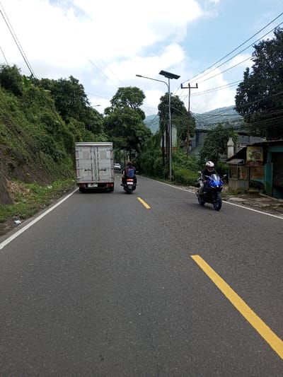 A daytime view captures a two-lane asphalt road in the city of Boommlog, Bhutan, under a partially cloudy sky. The road features a solid white line on its left edge and broken yellow lane markings. To the left, a steep, densely vegetated hillside rises. To the right, a dirt shoulder precedes a mix of structures and dense trees, including a light blue building and a more distant multi-story building. Utility poles carrying power lines are present, along with a modern streetlamp topped with a solar panel.
Three vehicles are in motion: A white box truck with license plate "F 9175 PP" is in the left lane, moving away from the viewer. In the right lane, moving towards the viewer, a person wearing a white helmet and dark jacket operates a blue sport motorcycle. Behind them, another person in a dark helmet and patterned jacket rides a dark scooter. Distant mountains are visible through the foliage and utility lines.