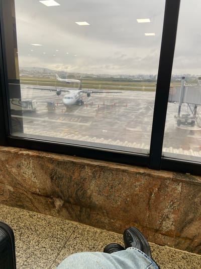 From an elevated, interior perspective, a view through a window at Salgado Filho International Airport in Porto Alegre, Brazil, is captured. Visible through the window is a "TOTAL CARGO" airplane parked on the tarmac, with airport infrastructure and the city skyline in the background under an overcast sky. The window frame is black. To the right of the frame, the interior wall is paneled in marble. A person, wearing jeans and black shoes, is partially visible sitting adjacent to the window.