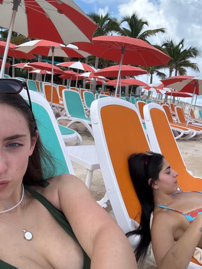 A daytime scene depicts a beach in the Berry Islands, Bahamas, with two women relaxing among rows of sun loungers and umbrellas. In the foreground, the left side of a woman's face, upper torso, and arm are visible; she has dark hair, blue eyes, and wears dark sunglasses on her head, a dark green bikini top, a white beaded necklace, and a silver locket. Her lips are slightly pursed, and she appears to be looking towards the camera while seated in a teal and white sun lounger. To her right, a second woman is shown in profile, facing right, with long dark hair, sunglasses on her head, and wearing a multi-colored bikini top. Her eyes are closed as she rests in an orange and white sun lounger. A small white plastic table stands between the two loungers. The background features numerous unoccupied sun loungers with white frames and alternating teal or orange fabric, arranged on light-colored sand. Many open beach umbrellas, primarily solid red or red and white striped, are scattered throughout the scene, providing shade. Palm trees are visible in the distant background under a partially cloudy sky.