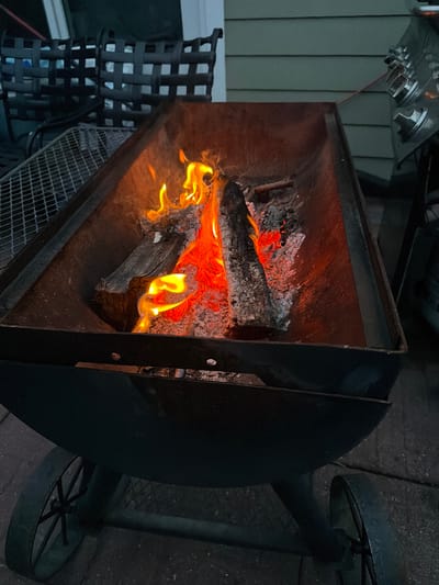 The image depicts a metal fire pit, likely a barbecue grill, containing burning logs and embers. The fire is active with visible flames and glowing coals. The fire pit is positioned outdoors on a paved surface, possibly a patio or driveway. In the background, there are glimpses of outdoor furniture, a house with siding, and what appears to be a propane tank. The overall scene suggests a backyard setting, potentially in Riverhead, United States of America, where outdoor cooking or a bonfire is taking place. The lighting suggests it is either dusk or a dimly lit evening.