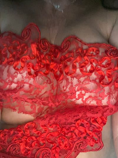 The image displays a close-up, overhead view of a person wearing red lace lingerie. The fabric is intricately detailed with floral patterns and swirling ribbon-like embellishments. The lighting is dim, creating a shadowed effect on the skin visible beneath the sheer lace. The background is dark and out of focus. There is no information in the image to suggest a location in Novi Petrivtsi, Ukraine.