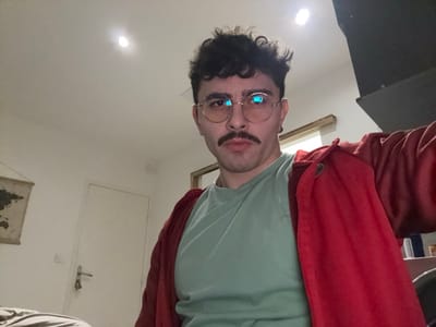 A man with curly dark hair, a mustache, and round glasses is angled slightly upward at the camera. He wears a light green t-shirt and a red jacket. A map hangs on the wall behind him to the left. There is a white door to the left of the frame. A dark object can be seen behind him to the right. The room is lit by ceiling lights.