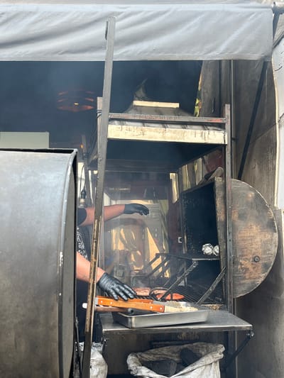 The image depicts a person wearing black gloves inside what appears to be a large metal barbecue smoker or grill. The person's arms are visible, reaching into the smoker. Several cooking tools, including knives with wooden handles, are visible, resting on a metal surface. The interior of the smoker is dark and greasy, with some aluminum foil balls visible on a rack. There is smoke present, indicating the smoker is in use. The exterior of the smoker appears weathered and used. A grey cloth or tarp is partially visible on the right. The environment appears to be an outdoor cooking setup, likely a food stall or catering operation. Details to suggest the location is San Francisco Totimehuacán, Mexico are not discernable in the image.