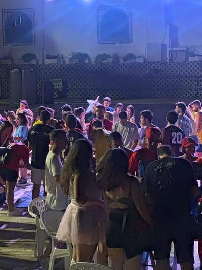 The image depicts an outdoor gathering of young adults at night in Blanca, Spain. The scene is illuminated by blue stage lighting, creating a vibrant and somewhat hazy atmosphere. A crowd of approximately 30-40 individuals, dressed in casual attire, are seen interacting, with many standing and conversing. Some individuals appear to be holding drinks. In the background, there is a wall with decorative lattice work and planters, suggesting an outdoor patio or courtyard setting. Two large speaker cabinets are visible on the left side of the frame. The overall impression is of a social event, possibly a party or a small concert.