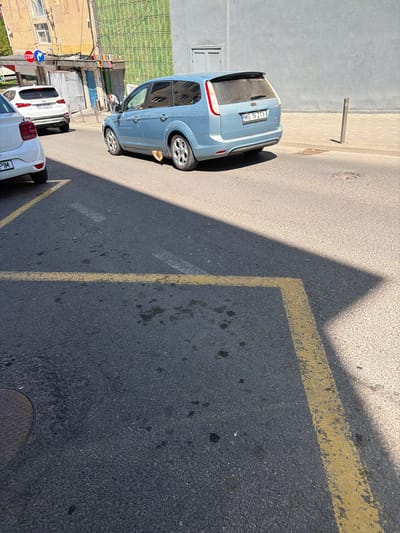 The image shows a street scene in Târgu Mureș, Romania. A light blue hatchback car with the license plate "MS 19 ZIX" is parked beside a sidewalk. There is a yellow parking line and triangle shadow on the road. There are two additional cars further down the road. There is a building on the left side. In the distance are street signs and an intersection.