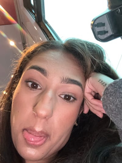 A selfie captures a woman with dark hair and brown eyes. Her hand rests on her forehead, partially obscuring a '1983' tattoo. She has a nose ring and her tongue is slightly visible between her lips. Natural light illuminates her face, highlighting her makeup, including well-defined eyebrows and blush. The background features a car's interior, including the sunroof and rearview mirror. A prism light reflection adds a vibrant touch to the image.
