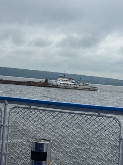 A barge is being pushed by a tugboat on the Volga River near the village of Yashel'cha, Russia. The sky is overcast, with clouds dominating the upper portion of the frame. The water is choppy, indicating a moderate breeze. The barge is laden with a dark-colored cargo, possibly soil or gravel, and appears to be quite long. The tugboat is white with blue accents and has a multi-decked superstructure. In the foreground, a metal railing with a blue top rail separates the viewer from the water. A mesh fence is attached to the railing, and a cylindrical metal receptacle, possibly an ashtray or trash can, is mounted on the fence. The far bank of the river is lined with a dense, green forest. The overall scene suggests a working river landscape on a cloudy day.