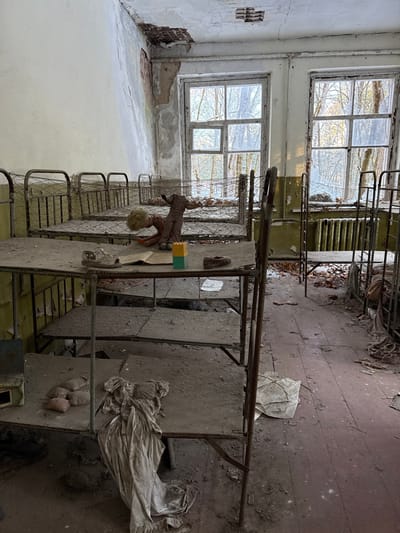 An abandoned room features rows of dilapidated metal bunk beds, coated in dust and debris. Two large windows illuminate the space, showcasing cracked walls and peeling paint. A doll rests on one bed, adding an unsettling element. Discarded materials and remnants of past occupants litter the floor. The room exudes a sense of decay and neglect.
