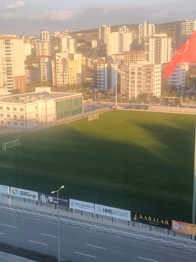 The image captures a high-angle urban landscape, likely in Etimesgut, Turkey, bathed in the warm, golden light of either late afternoon or early morning.

In the foreground, a multi-lane asphalt road is visible, demarcated by white lines, with a solitary street light positioned centrally. Below the football field, behind a dark green fence, a series of advertising billboards are displayed. The legible text on these billboards includes "SERGrup", "Etimesgut'un sağlığı", "KEKLİKOĞLU GRUP", "HMS İNŞAAT", "KARALAR İNŞAAT", and "Ayber İNŞAAT & MÜHENDİSLİK".

The midground is dominated by a meticulously kept green football (soccer) field, complete with white line markings for the penalty areas, center circle, and halfway line. Two white goalposts stand ready at opposite ends of the pitch. Adjacent to the field, on the left, is a modern multi-story building featuring a light exterior and prominent green-tinted windows, with parts of its roof showing HVAC units. Long shadows stretch across the field, a notable detail indicating the low angle of the sun. No activity or people are visible on the pitch, suggesting a quiet moment.

The background is filled with a dense array of residential high-rise buildings, varying in design and height, many reflecting the golden sunlight. These structures ascend towards distant, low-lying hills covered with trees and sparse vegetation, defining the city's edge. A vibrant red Turkish flag unfurls prominently on the right side of the frame, adding a national emblem to the scene. Another sign visible in the mid-background reads "ARMONI PARK".

The scene conveys the development and structure of a modern city, with the football field providing a green recreational contrast amidst the surrounding buildings. The clear sky and soft, warm lighting contribute to a tranquil atmosphere.