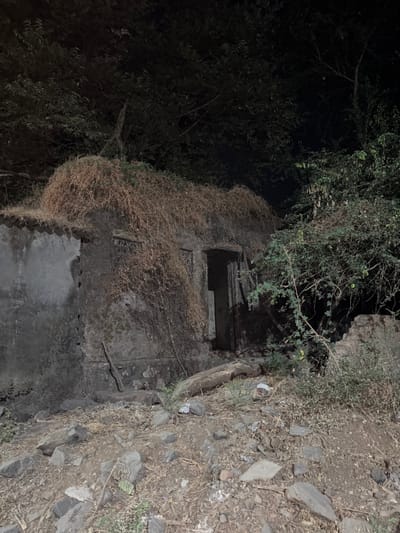 The image displays a dilapidated single-story structure at night, located in Ambernath, India. The building's exterior walls are made of concrete or stone, exhibiting significant weathering, discoloration, and moss growth. A substantial portion of the roof and the upper facade, particularly on the right, is covered with a dense layer of dry, light brown, stringy vegetative matter. The structure features a central, dark, rectangular doorway with a visible frame; the interior beyond is obscured. Two apparent window openings are visible on the front wall, one to the left and one to the right of the doorway, both appearing dark and potentially boarded or grilled. The ground in the foreground is uneven, covered with loose soil, various sizes of grey rocks, dry leaves, and sparse green plant growth. Minor debris, including what appears to be discarded paper or plastic, is scattered near the base of the structure, alongside a horizontal log or thick branch positioned near the doorway. Dense, dark green foliage from trees and bushes heavily surrounds the building, occupying the background and right side of the frame. The overall scene is dimly lit under a dark sky, indicating nighttime conditions. No individuals, actions, or interactions are present in the image.