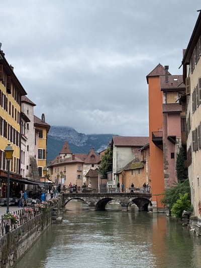 The image shows a view of the Thiou River in Annecy, France, with its canalized section flowing through the historic old town. On the left, a row of multi-story buildings with yellow and pale pink facades line the riverbank. These buildings feature traditional architecture with multiple windows and shutters, and a sidewalk with outdoor seating for a cafe is visible at street level. Streetlamps with ornate designs are positioned along the sidewalk.

A stone bridge with three arches spans the river, connecting the two sides of the town. People are walking across the bridge. On the right side of the river, more buildings of varying colors and architectural styles are visible, including an orange building with a small balcony and a tall, narrow pink building. Lush green foliage cascades down some of the walls.

In the background, a mountain range is shrouded in clouds, creating a dramatic backdrop. The sky is overcast, suggesting a cloudy or possibly rainy day. The river itself is calm, reflecting the overcast sky and the surrounding buildings. Several individuals are visible in various locations: on the sidewalk, on the bridge, and near the river's edge. Their actions are casual, indicative of daily life in the city. The overall scene captures the picturesque charm of Annecy's waterways and historic architecture.