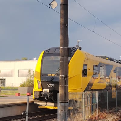 The image shows the front of a yellow and black train stopped at a station. The train has the number "16EV-0013" printed on its front. To the left of the train is a building with large windows, and to the right is a fence and some dry vegetation. A utility pole stands in front of the train. The sky is cloudy. The location is the Jelgava train station in Jelgava, Latvia.