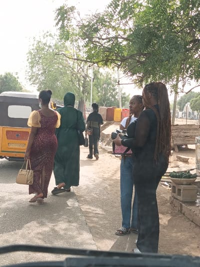Four women and a man are on a street. One woman is in a burgundy dress with sheer sleeves, carrying a beige purse. Another is wearing a dark green robe. A man walks behind them, and two women in modern clothing stand on the right. One wears jeans and a white top, while the other has on a black outfit with long hair. A yellow vehicle sits to the left, and colorful containers line the background. A small fruit stand is on the right side of the road. The street scene is shaded by trees.