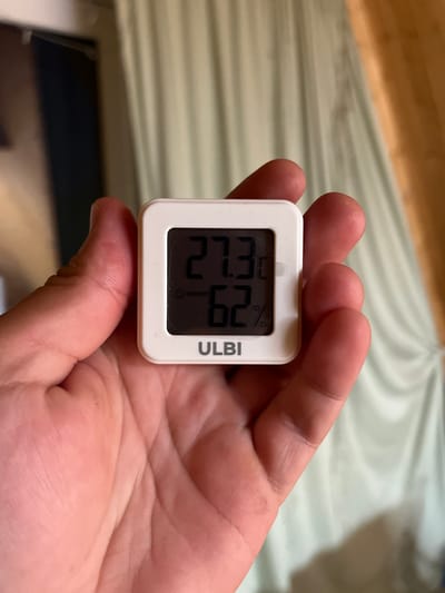 A person is holding a white digital thermometer in their right hand, with their palm and fingers visible. The thermometer displays a temperature of 27.3°C and a humidity reading of 62%. The background consists of light-colored curtains. The image's metadata does not contain any information about the location of Vasilyevo, Russia, or any other geographical details.