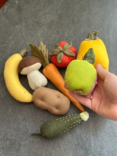 A flat lay of handmade fabric toy vegetables and fruit is displayed on a dark gray textured surface, likely a countertop or table. The collection includes a yellow banana, a brown and white mushroom, a brown potato, an orange carrot, a red tomato, a yellow pumpkin, a green pickle, and a green apple. A person's hand, wearing a gold ring on the right ring finger, is visible on the right side of the image, holding the green apple. The background is a plain, dark gray surface with a subtle texture. The scene is lit from above, casting soft shadows. The image is an artistic arrangement of fabric toys, with no indication of location in Chornomorske, Ukraine.