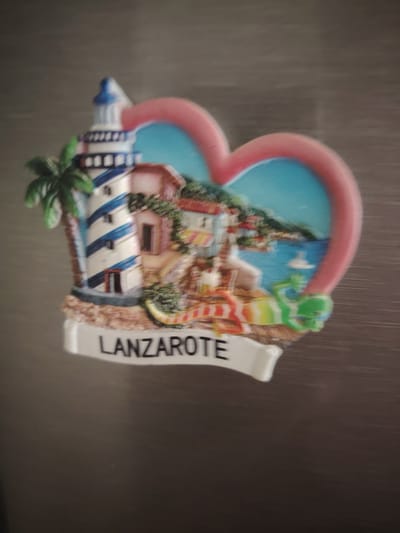 A heart-shaped magnet is attached to a metallic surface, likely a refrigerator. The magnet depicts a scene with a lighthouse, buildings, a palm tree, and a view of the sea with rolling hills in the background. A small green figure, possibly a lizard or frog, is positioned near the water. The word "LANZAROTE" is written in black capital letters on a white banner at the bottom of the magnet. The background is a blurred, neutral gray surface. The image quality is poor due to blurriness and low resolution. There is no information in the image to suggest a location in Huntingdonshire, United Kingdom.