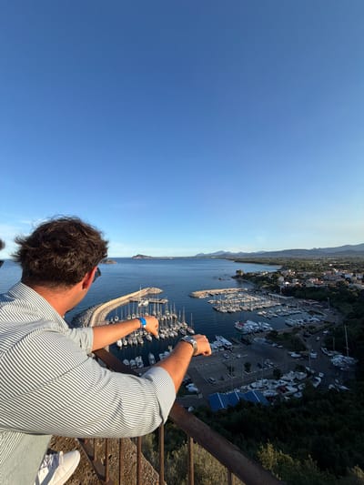 An elevated, over-the-shoulder perspective looks out from a balcony at a man facing away from the camera, his head and shoulders in the foreground. He wears a light-colored, horizontally striped shirt and white sneakers. His right arm is extended, resting on a rusty railing. The man is looking towards a large harbor filled with numerous sailboats and smaller vessels.

The harbor is situated in Santa Maria Navarrese, Italy, and is surrounded by a coastal town with buildings clustered along the shoreline and extending inland. A long, curved breakwater protects the harbor. Beyond the town and harbor, a verdant landscape with hills and mountains forms the backdrop under a clear, pale blue sky. The scene is bathed in the warm light of late afternoon or early evening.