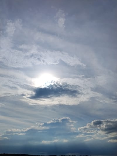 This media file captures a dramatic sky scene with the sun peeking through a break in the clouds. The sky is filled with a variety of cloud formations, ranging from thin, wispy cirrus clouds stretching across the upper portion of the frame to darker, more substantial cumulus clouds lower down. The sun, a bright, almost blinding orb, is partially obscured by a thicker cloud, creating a halo effect and casting rays of light that pierce through the atmospheric haze.

The overall mood is one of dynamic weather, perhaps hinting at an approaching or receding storm, or simply a lively atmospheric condition. The lighting suggests it is either sunrise or sunset, given the intensity of the sun and the way it interacts with the clouds. There are no people or identifiable objects in the image, focusing entirely on the celestial display. The bottom of the frame shows a dark, silhouetted horizon, possibly indicating land or distant structures, but these are indistinct.

The location is Ustyvytsya, Ukraine. The weather appears to be partly cloudy with some dramatic cloud formations and bright sunlight. The emotional tone conveyed is one of awe and the power of nature. There is no visible text in the image.