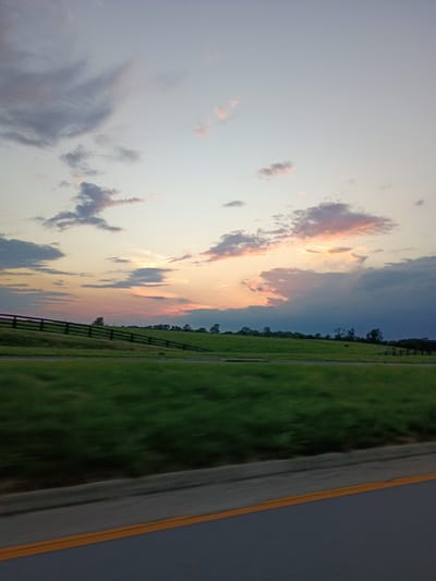 An outdoor, vertical, eye-level shot depicts a tranquil countryside scene at dusk. The sky transitions from a pale blue to shades of orange and pink, with scattered clouds. A dark, wooden fence runs horizontally across the middle ground, separating a lush green field from the sky. The field is slightly blurred, suggesting motion, and stretches towards a tree line in the distance. The foreground shows a road with a yellow line, also blurred. The overall atmosphere is peaceful and serene, with no people or animals visible. The image was captured in Lancaster, United States.