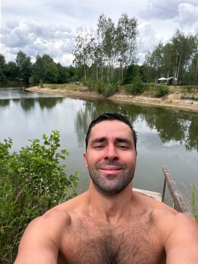 An outdoor, selfie-style photograph shows a shirtless man with dark hair and a beard standing on a wooden dock. The man is smiling and looking directly at the camera. The background features a body of water, a sandy shore, and a line of trees, including birch trees, under a cloudy sky. In the distance, there are some structures, including an umbrella and what appears to be a small building. The man's chest and shoulders are visible, showing dark body hair. The water reflects the trees and the sky. The overall setting suggests a natural environment, possibly a lake or pond, within the vicinity of Dobrzyń, Poland.