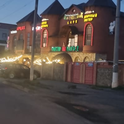 A twilight exterior view shows a brick building with multiple towers and arched windows, illuminated by neon signs and string lights. The main facade displays several signs, including "FAMILY CAFE" in yellow and green lettering, and further to the left, red neon letters spelling "АРЦАХ". To the right, yellow neon letters form the words "ШАШЛЫЧНЫЙ ПАРТНР". Below these signs, a horizontal green neon sign reads "ТРАКТОР НА МАНГАЛЕ".
In front of the building, a dark-colored car is parked on the street. The building is set back from the road, with a decorative red fence to the right. The environment suggests a commercial establishment, possibly a restaurant or cafe, in the city of Aktobe, Kazakhstan. The lighting indicates it is evening or dusk. No people are clearly visible in the image.