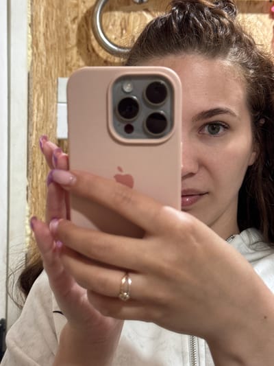 A woman is taking a selfie with a pink iPhone. She is wearing a white shirt. She has a ring on her finger. The location appears to be a bathroom, with a wooden wall and white trim. The location of Nizhnyaya Gar', Russia cannot be verified from the image.